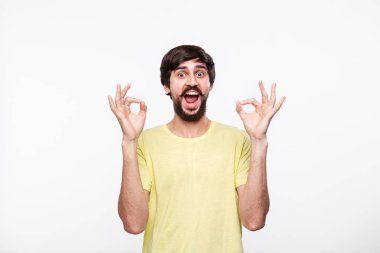 Yakışıklı brunet sakallı adam sarı gömlek bıyıklı ok işareti gösteren iki eli beyaz arka plan üzerinde izole ayakta. Tamam hareketi.