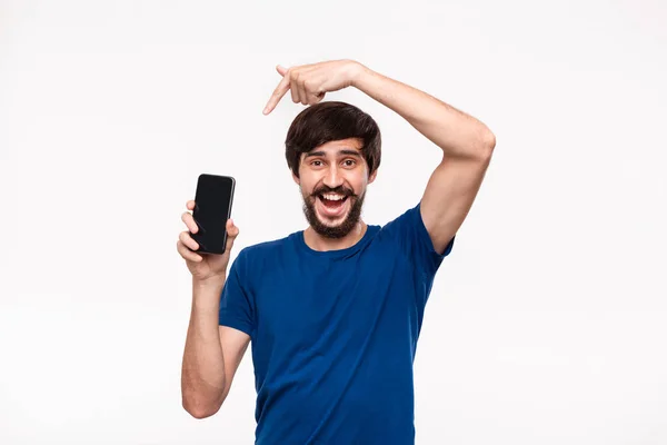 Good looking brunet man in a blue shirt with beard and mostaches excited looking at the camera pointing at the smartphone standing isolated over white background. Emotion and gesture of surprise. — Stock Photo, Image