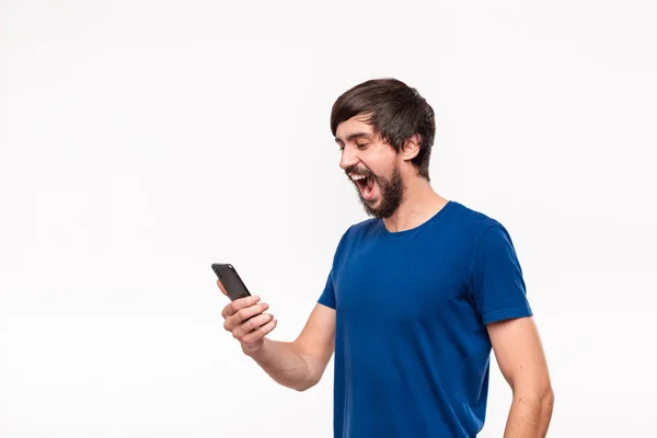 Handsome brunet man in a blue shirt with beard and mostaches surprised holding smartphone looking at the camera standing isolated over white background. Emotion and gesture of surprise. — Stock Photo, Image