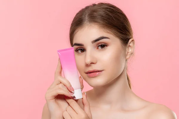 Güzel kız cildi nemlendirmek için krem tüp tutan. Model pembe arka plan üzerinde izole kamera bakar. Güzellik ve sağlık tedavisi kavramı.