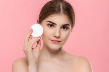 Güzel model beyaz pamuk sünger ile cilde kozmetik ürün uygular. Model pembe arka plan üzerinde izole kamera bakar. Güzellik ve sağlık tedavisi kavramı.