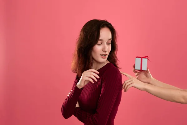 Happy pretty brunette girl gets a present box with a ribbon from another person standing isolated on a dark pink background and smiling at the camera. Celebrating special event. — Stock Photo, Image