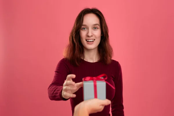 Feliz chica morena bonita recibe una caja de regalo con una cinta de otra persona de pie aislado sobre un fondo de color rosa oscuro y sonriendo a la cámara. Celebración de un evento especial . —  Fotos de Stock