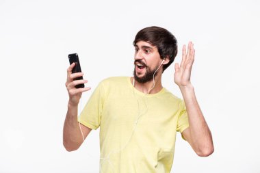Bıyıklı yakışıklı brunet sakallı adam beyaz arka plan üzerinde izole bir akıllı telefon ile arkadaşlarınızla sohbet video. Model ses için beyaz kablolu kulaklık kullanır.