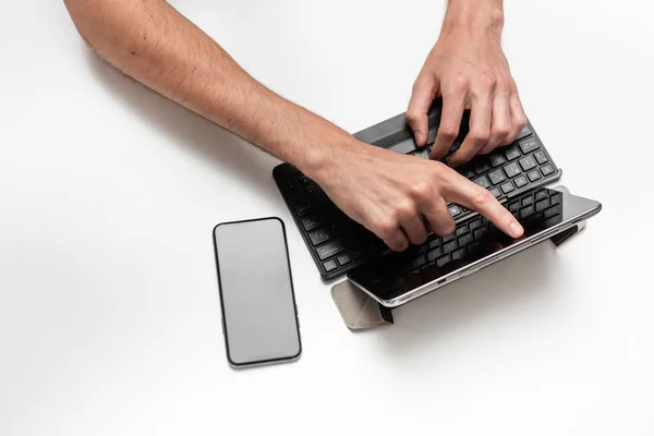 Close up top view of a man working on a project using digital tablet with keyboard seating at white table. Smartphone is near the tablet. Concept of using modern technologies at work.