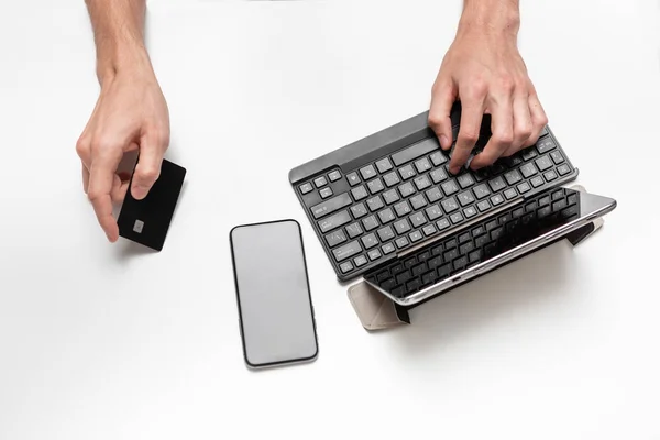 Close up top view of a man working on a project using digital tablet with keyboard seating at white table. Smartphone and bank card is near the tablet. Concept of using modern technologies at work.