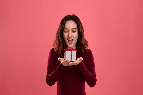 Vzrušená pěkná brunetka, která drží tu skříňku s stuhou se dvěma rukama na tmavém růžovém pozadí a usmívajíje se v kameře. Oslava zvláštní události. — Stock fotografie