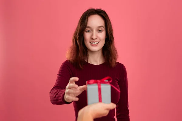 Szczęśliwa Ładna brunetka dostaje obecne pudełko z wstążką z innej osoby stojący izolowane na ciemnym różowym tle i uśmiechając się do aparatu. Uroczysta impreza specjalna. — Zdjęcie stockowe