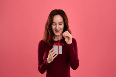 Excited pretty brunette girl opens gift packed in present box with a ribbon standing isolated on a dark pink background. Celebrating special event.