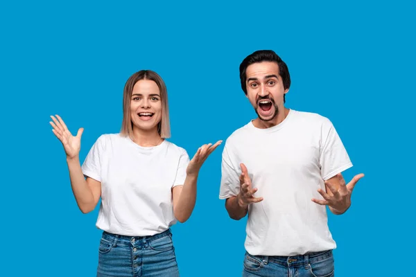 Pareja feliz expresando emoción de éxito. Joven mujer rubia y hombre barbudo con bigotes en camisetas blancas y pantalones vaqueros azules son los ganadores. Modelos de pie aislados sobre fondo azul . —  Fotos de Stock