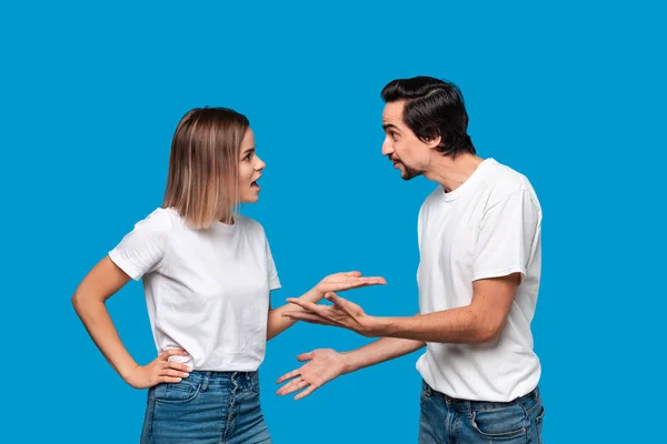 Couple of a young blond woman and brunet bearded man with mustaches in white t-shirts and blue jeans quarreling standing isolated over blue background.