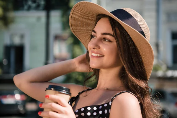 Chica bonita en un vestido negro y sombrero con solapas anchas sostiene la taza de papel desechable de café caliente disfrutando de caminar por la ciudad europea durante el soleado día de verano. Viajar en verano — Foto de Stock