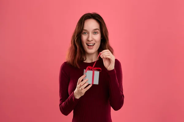 Emocionado chica morena bonita abre regalo embalado en la caja actual con una cinta de pie aislado sobre un fondo de color rosa oscuro y sonriendo a la cámara. Celebración de un evento especial . —  Fotos de Stock