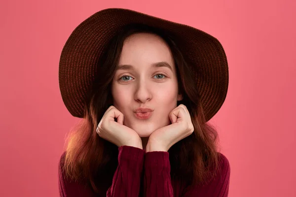 Emocionado hermosa chica morena en un sombrero de ala ancha enviando besos de aire de pie sobre un fondo rosa oscuro . —  Fotos de Stock