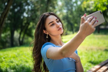 Mavi elbiseli gülümseyen genç kadın parkta oturuyor ve akıllı telefonla selfie çekiyor.