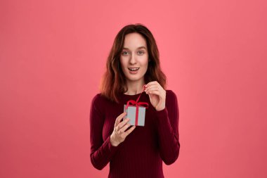 Gülümseyen güzel esmer kız hediye koyu pembe arka plan üzerinde izole ayakta ve kameraya gülümseyerek bir kurdele ile mevcut bir kutu içinde paketlenmiş açılır. Özel etkinliği kutluyoruz.