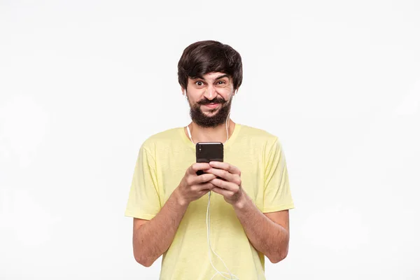 Brunet bearded man with whiskers in a yellow t-shirt holding smartphone with connected wired headphones expressing emotion of distrust isolated over white background. Receiving unexpected news. — Stock Photo, Image
