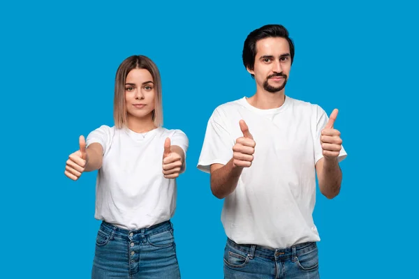 Hombre barbudo joven feliz con bigotes y mujer rubia en camisetas blancas y pantalones vaqueros con pulgares arriba de pie aislado sobre fondo azul. Concepto de evaluación pisiva . — Foto de Stock