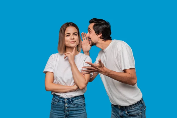 Bearded man with mustaches in white t-shirt tells secret to a young blond girl in white t-shirts and blue jeans standing isolated over blue background. Concept of secret telling.