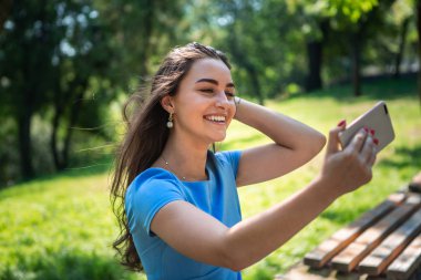 Mavi elbiseli gülümseyen genç kadın parkta oturuyor ve akıllı telefonla selfie çekiyor.
