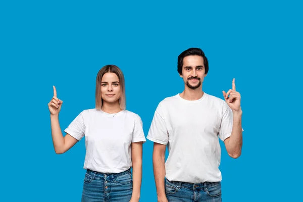Couple souriant en t-shirts et jeans blancs pointant vers le haut avec les doigts isolés sur fond bleu. Espace de copie pour la publicité produit . — Photo