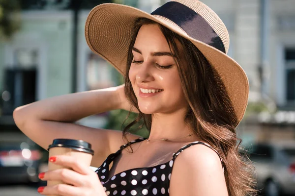 Chica bonita en un vestido negro y sombrero con solapas anchas sostiene la taza de papel desechable de café caliente disfrutando de caminar por la ciudad europea durante el soleado día de verano. Viajar en verano — Foto de Stock