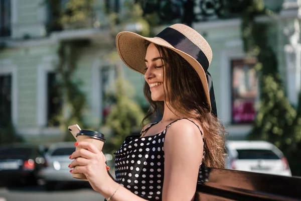Chica bonita en un vestido negro y sombrero con solapas anchas sostiene la taza de papel desechable de café caliente en una mano y teléfono inteligente en otro disfrutando de caminar durante el soleado día de verano . — Foto de Stock