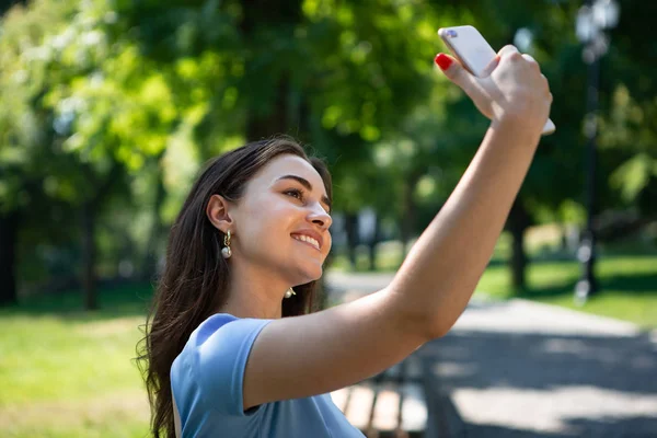 Mavi elbiseli gülümseyen genç kadın parkta oturuyor ve akıllı telefonla selfie çekiyor.
