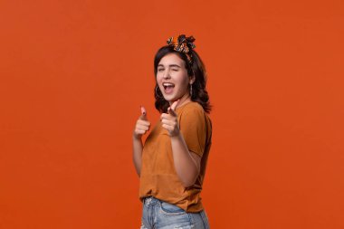 Bir t-shirt ve güzel kafa bandı turuncu arka plan üzerinde izole kamera parmaklarıyla işaret Güzel esmer kadın. Reklam yeri.