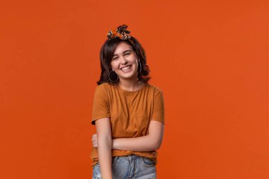 Turuncu bir t-shirt ve güzel kafa bandı turuncu arka plan üzerinde izole ayakta kameraya bakarak gülümseyen esmer kadın. 