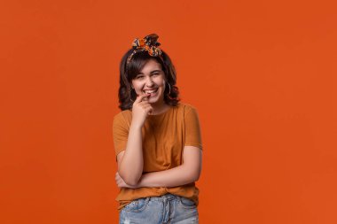 Turuncu bir t-shirt ve güzel kafa bandı turuncu arka plan üzerinde izole ayakta kameraya bakarak gülümseyen esmer kadın. 