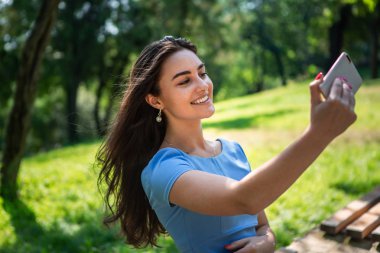 Mavi elbiseli gülümseyen genç kadın parkta oturuyor ve akıllı telefonla selfie çekiyor.