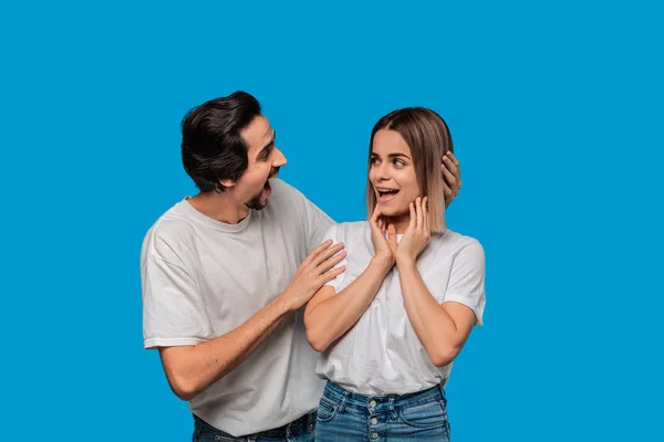 Happy couple of a young blond woman and brunet bearded man with mustaches in white t-shirts and blue jeans posing isolated over blue background. Concept of an ideal couple.