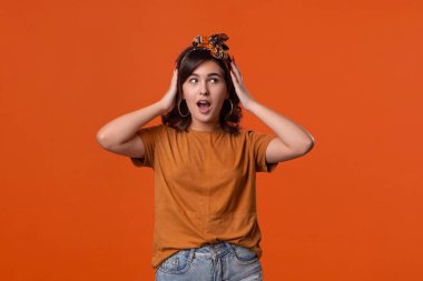 Bir t-shirt ve güzel kafa bandı turuncu arka plan üzerinde izole ayakta sürpriz jest ifade korkmuş esmer kadın. Reklam yeri.