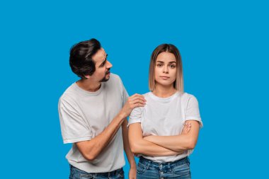 Brunet beyaz t-shirt ve mavi kot bıyıklı adam mavi arka plan üzerinde izole af için kız arkadaşı soran. Suçluluk kavramı.