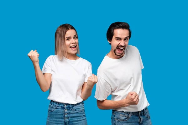 Emotional couple expressing gesture of success. Young blond woman and bearded man with mustaches in white tees and blue jeans are winner. Models standing isolated over blue background. Concept of succ