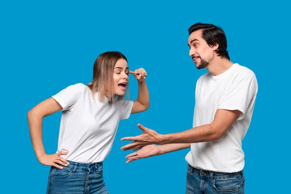 Eine junge blonde Frau und ein brünetter bärtiger Mann mit Schnurrbart in weißen T-Shirts und blauen Jeans streiten sich isoliert vor blauem Hintergrund. Konzept der Beziehungskrise. — Stockfoto