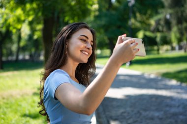 Mavi elbiseli gülümseyen genç kadın parkta oturuyor ve akıllı telefonla selfie çekiyor.