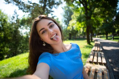 Mavi elbiseli gülümseyen genç kadın parkta oturuyor ve akıllı telefonla selfie çekiyor.