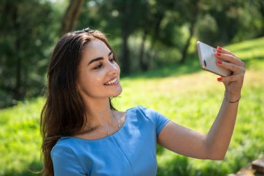 Mavi elbiseli gülümseyen genç kadın parkta oturuyor ve akıllı telefonla selfie çekiyor.