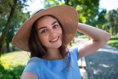 Geniş kapaklı mavi elbise ve şapkalı gülümseyen genç kadın parkta oturur ve akıllı telefonla selfie çeker.