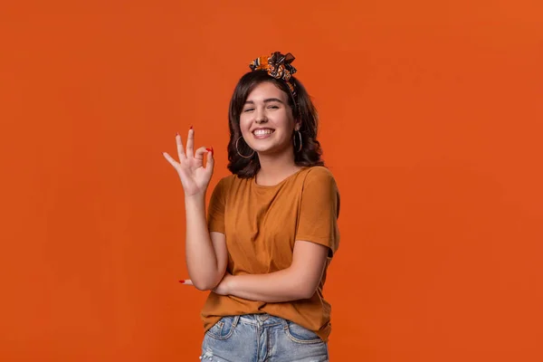 Smiling brunette woman in an orange t-shirt and beautiful headband showing OK-sign with fingers standing isolated over orange background. Concept of positive evaluation.