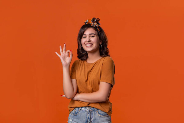 Smiling brunette woman in an orange t-shirt and beautiful headband showing OK-sign with fingers standing isolated over orange background. Concept of positive evaluation.