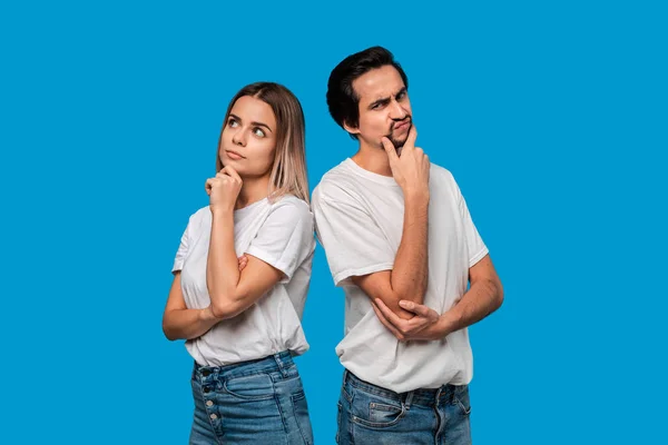 Thoughtful couple of a bearded brunet man with mustache and blond woman in white t-shirts and blue jeands standing isolated over white background.
