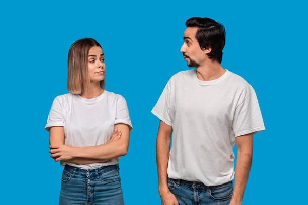 Couple d'une jeune femme blonde et d'un homme barbu brunet avec moustaches en t-shirts blancs et jeans bleus se regardant posant isolés sur fond bleu. Concept d'un couple idéal . — Photo