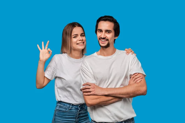 Happy couple of a young blond woman and brunet bearded man with mustaches in white t-shirts and blue jeans posing isolated over blue background. Concept of an ideal couple.