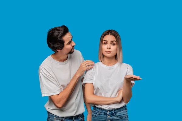 Brunet bearded man with mustaches in white t-shirt and blue jeans asking his girlfriend for forgiveness isolated over blue background. Concept of guilt.