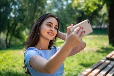 Mavi elbiseli gülümseyen genç kadın parkta oturuyor ve akıllı telefonla selfie çekiyor.