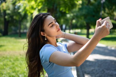 Mavi elbiseli gülümseyen genç kadın parkta oturuyor ve akıllı telefonla selfie çekiyor.
