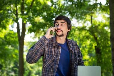 Sakallı ve bıyıklı genç bir esmer parkta dizüstü bilgisayarla oturuyor ve akıllı telefondan konuşuyor. Herhangi bir yerde çalış.